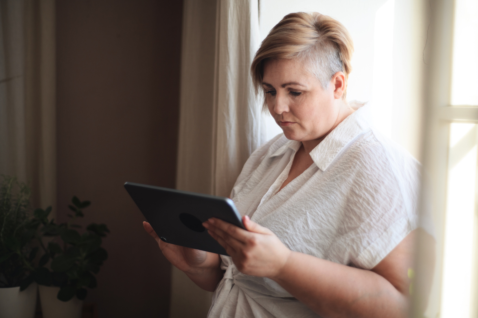 Woman in White Shirt Standing by Window and Using Tablet.